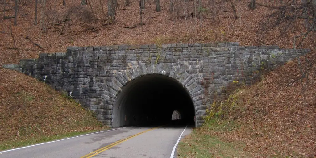 Stone tunnel entrance on the Blue Ridge Parkway, cut through a wooded hillside, with the tunnel's far end visible as a point of light.