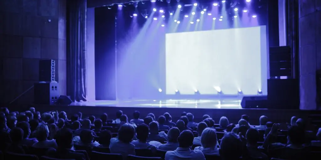 Audience seated in a darkened auditorium facing a brightly lit but empty stage with a blank screen.