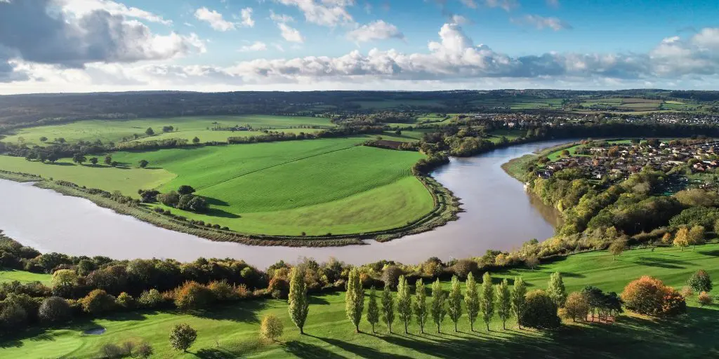 Aerial view of a wide river making a broad, calm bend around a green tongue of farmland, banked by trees on both sides, with a row of poplars in the foreground.