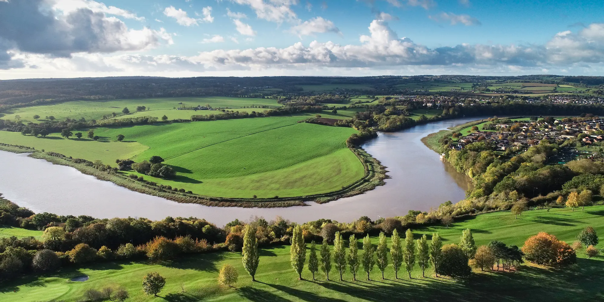 Aerial view of a wide river making a broad, calm bend around a green tongue of farmland, banked by trees on both sides, with a row of poplars in the foreground.