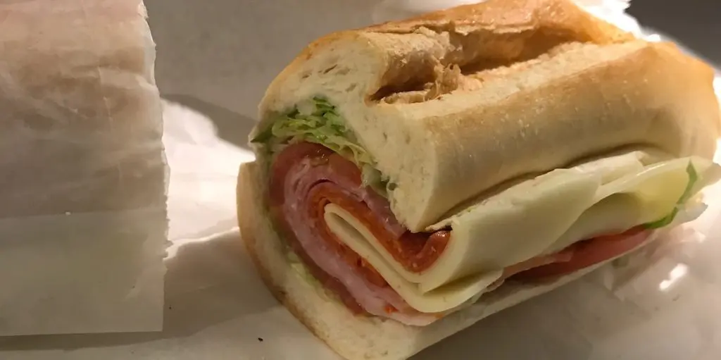 An Italian sub on a split-top roll, wrapped in deli paper, layered with ham, salami, pepperoni, provolone, and shredded lettuce.